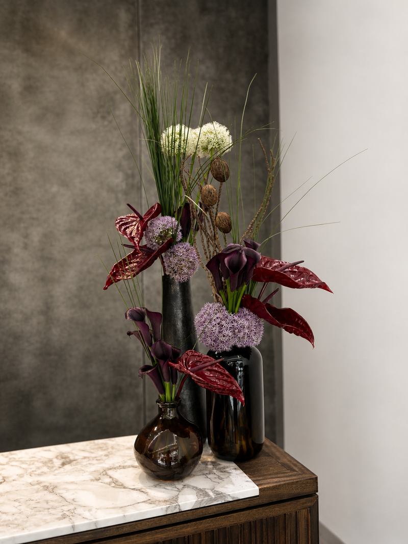 Decorative floral arrangement with purple and green elements in dark glass vases on a marble surface.